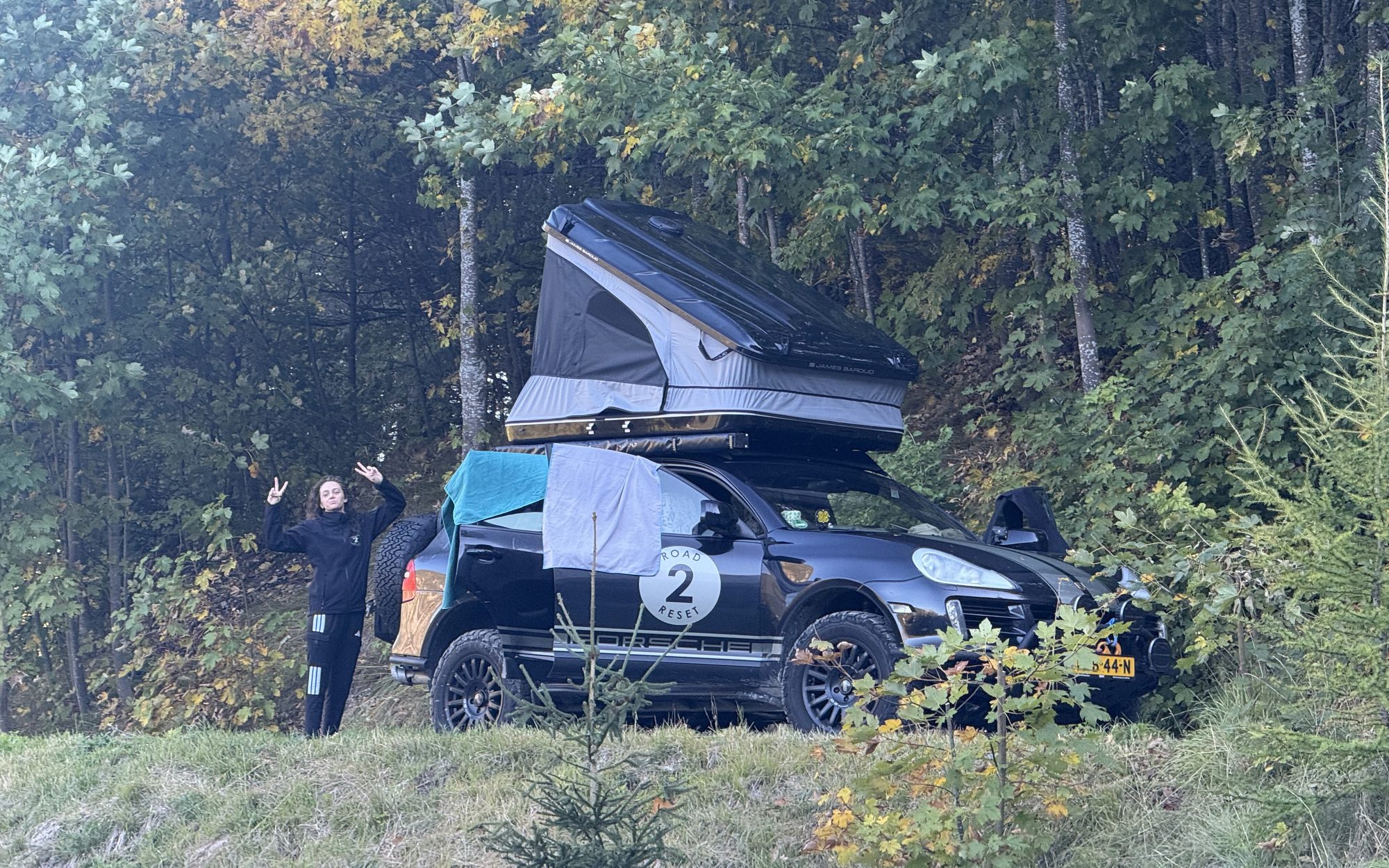 Tess making peace signs next to the Cayenne with the roof tent open and towels drying, at the edge of the forest