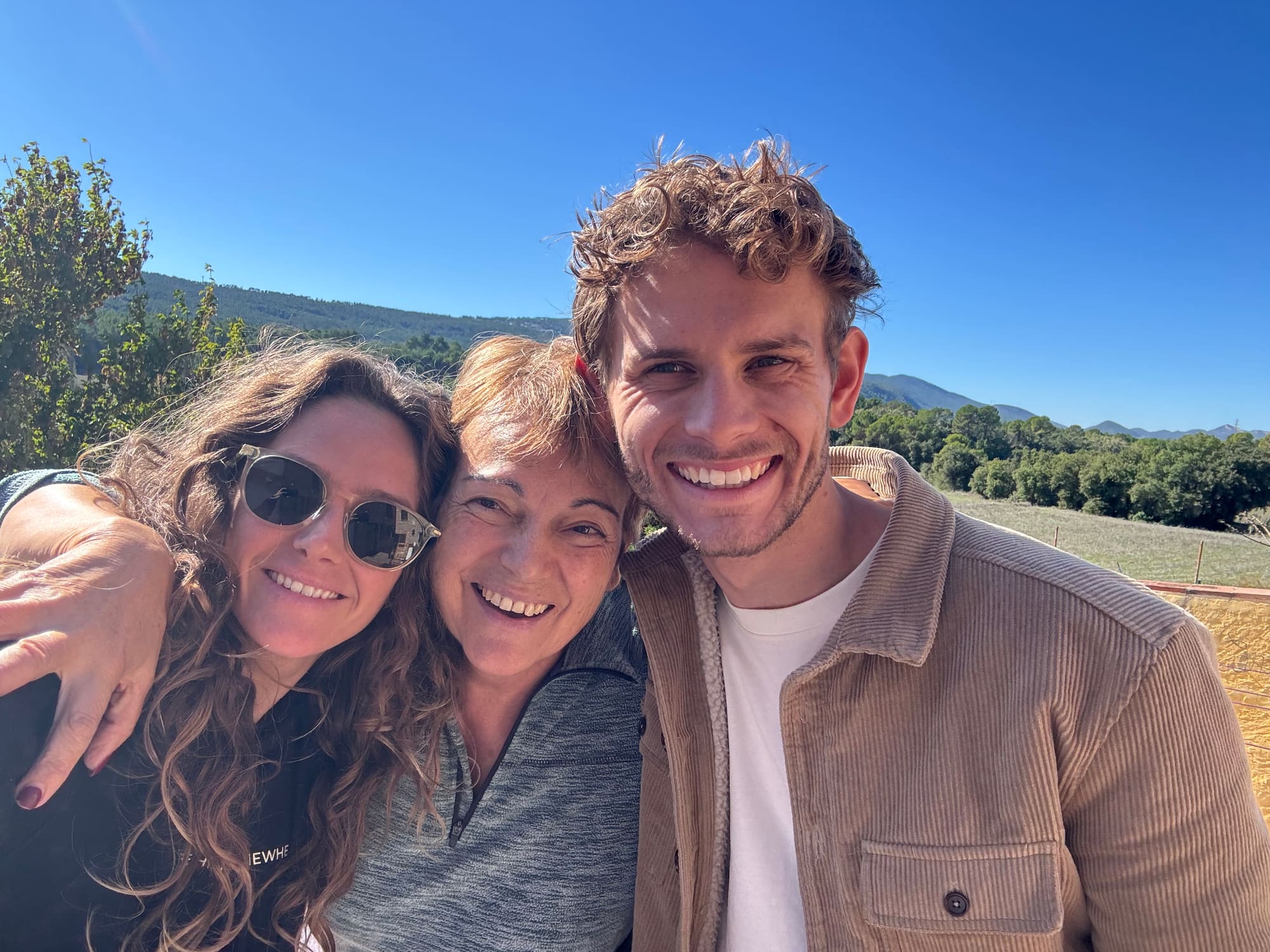 Tess, Bodhi and a woman between them smiling into the camera in the sunshine, with green hills and mountains under a blue sky behind them