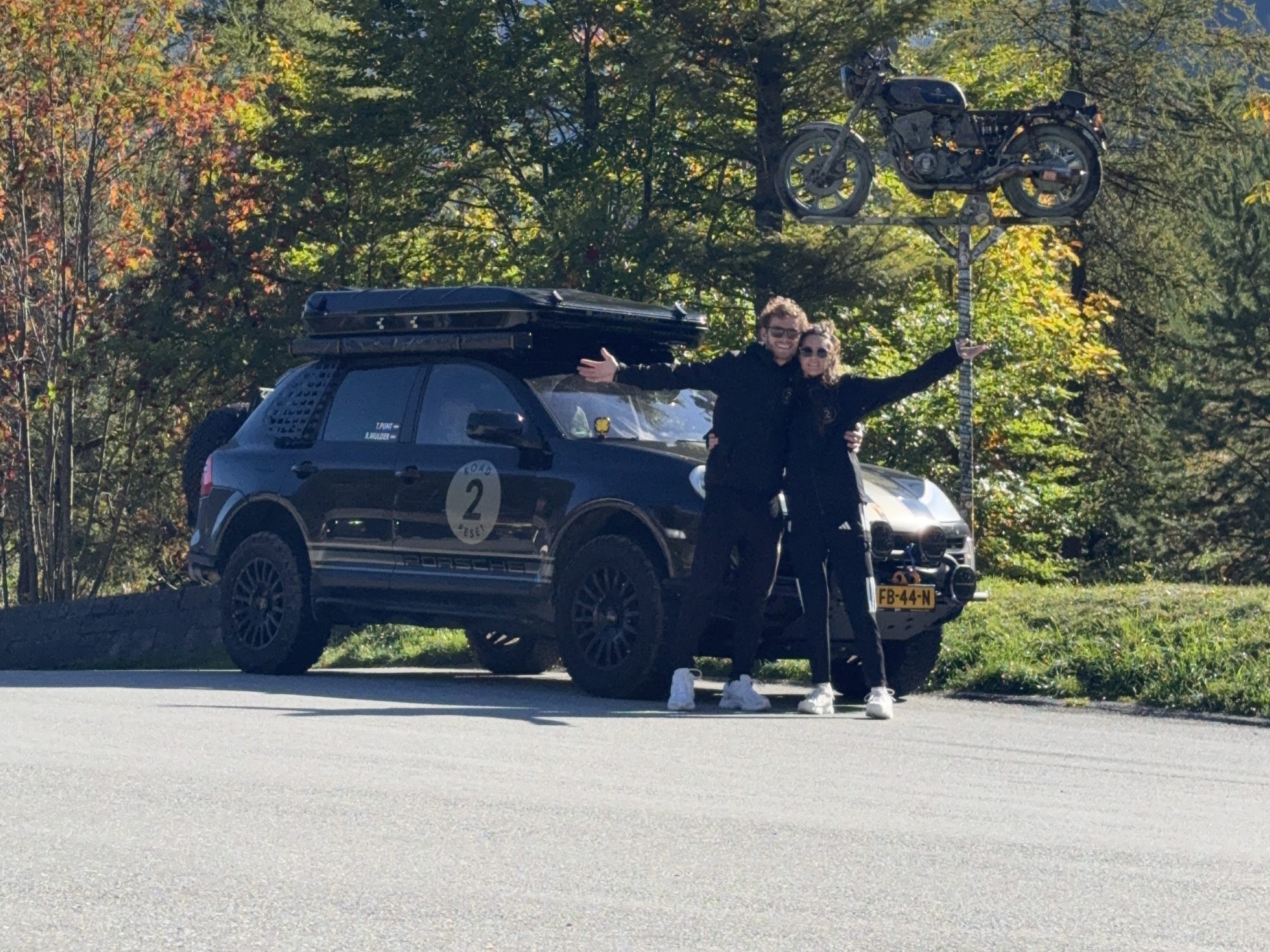 Bodhi and Tess with arms wide in front of the Cayenne, autumn trees and Alpine peaks behind them