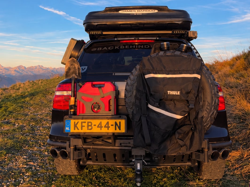 The back of our Porsche Cayenne on a grassy ridge at sunset, with jerry cans, a shovel, a red diesel can, the spare wheel in a Thule bag and mountains behind