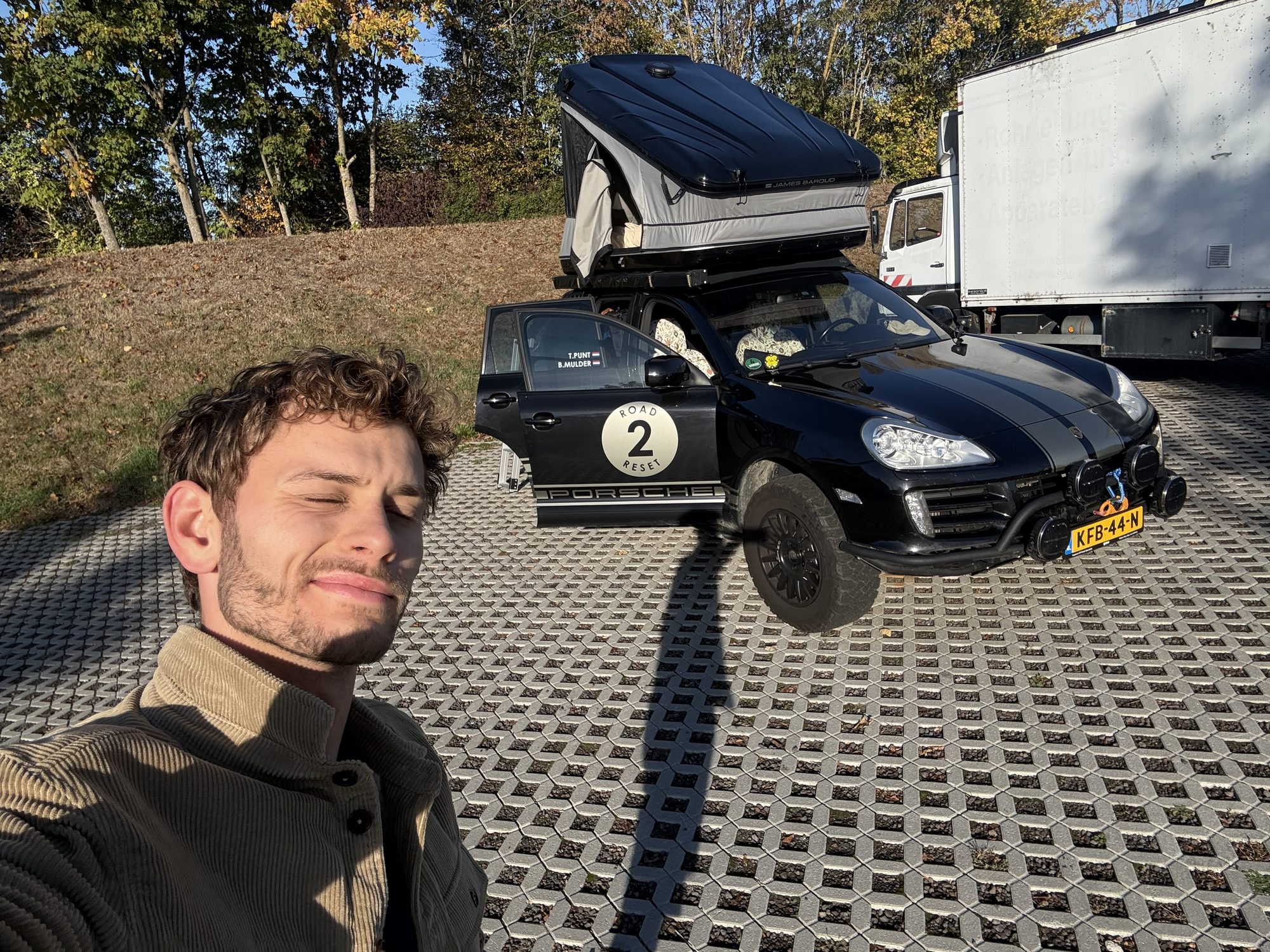 Bodhi squinting into the morning sun, the Cayenne behind him with the roof tent open on a car park near Mainz