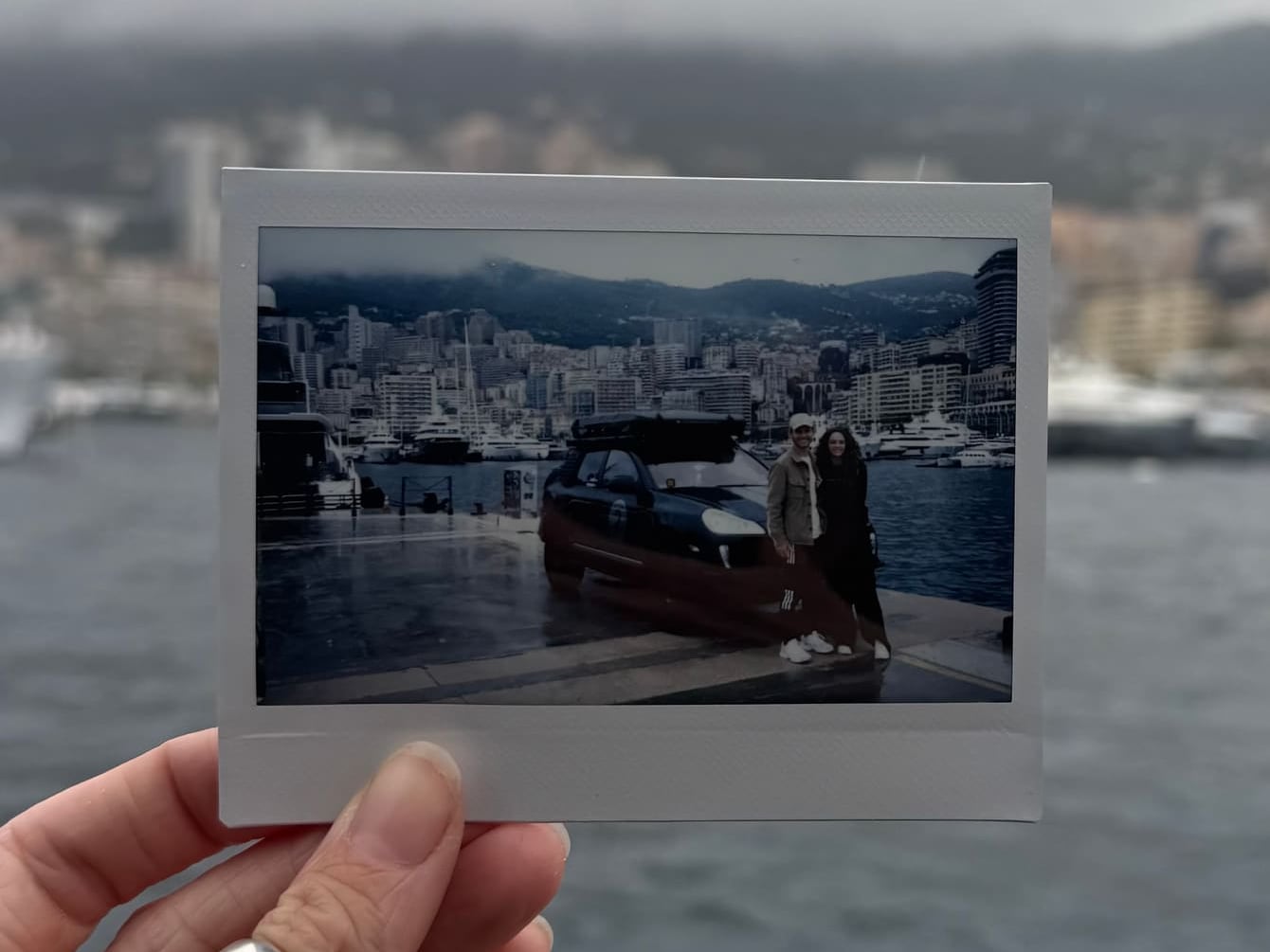 A hand holding up a polaroid of Bodhi and Tess with the Cayenne by the harbour in Monaco, the same harbour and the clouded hills blurred behind it