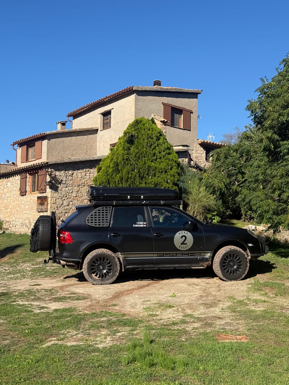 The Cayenne parked on the grass in front of a stone house with brown shutters, mud on the tyres, under a clear blue sky