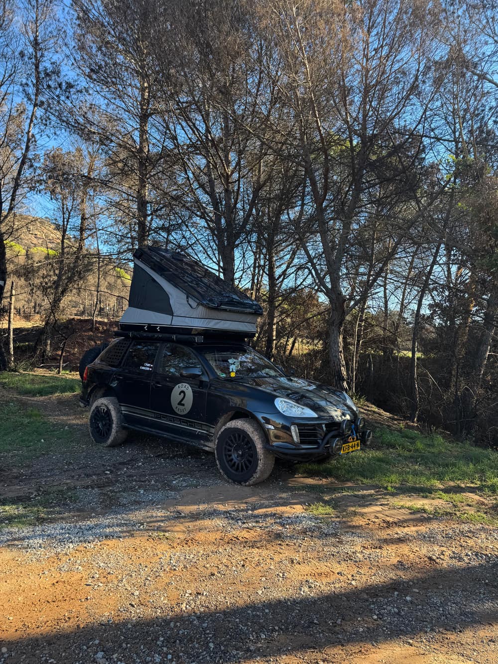 The Cayenne with its roof tent folded out on a gravel track among pine trees near Carme, mud on the tyres, in the low sun